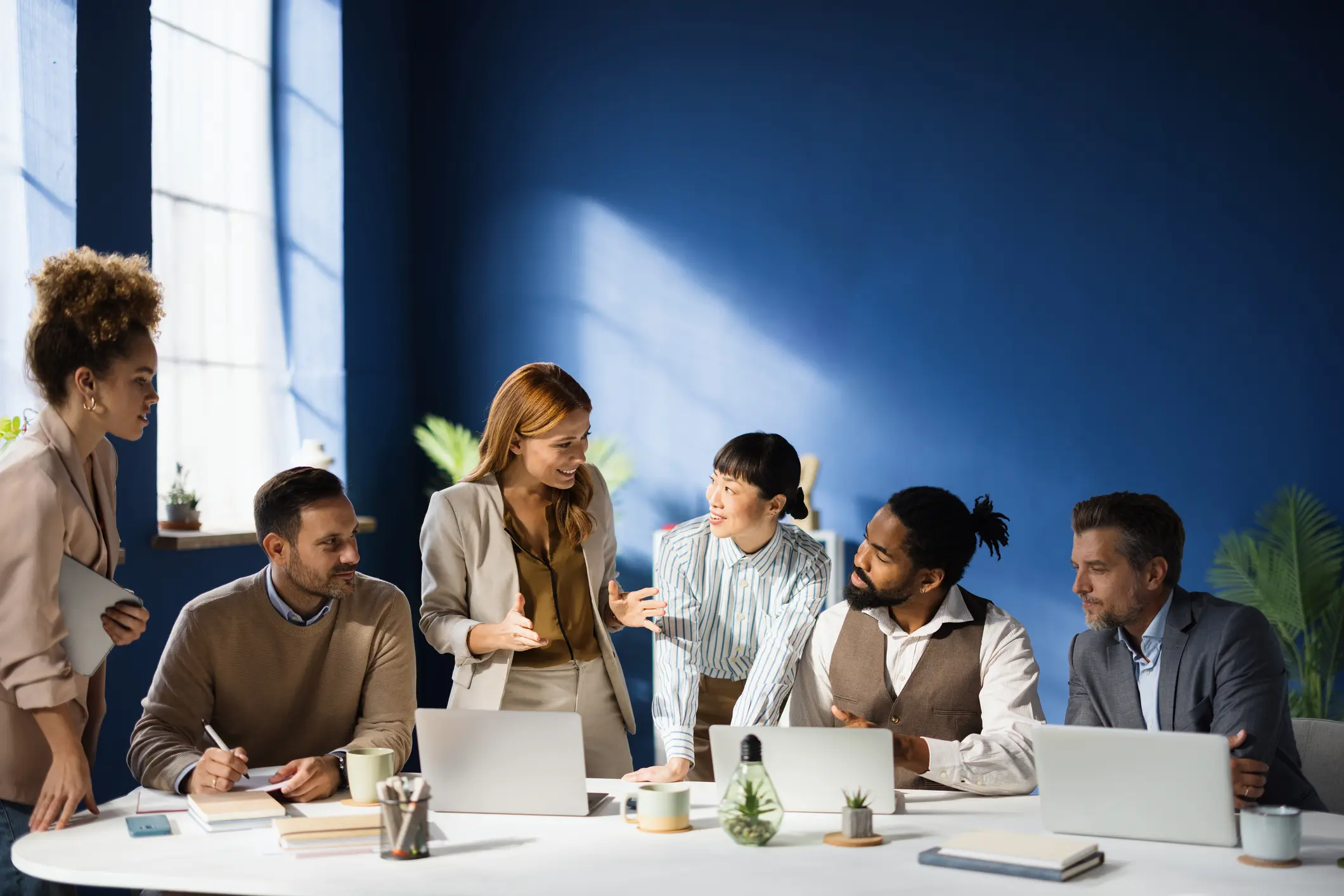 Diverse group of professionals engaging in a collaborative discussion around a table in a modern office setting. Laptops and plants create a productive atmosphere